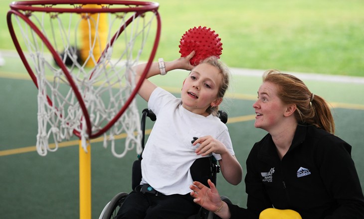 Girl in wheelchair shooting a basket as helper watches