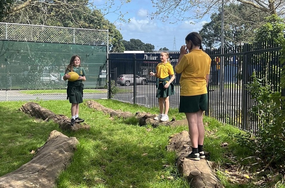 Three tamariki playing with a ball standing on logs
